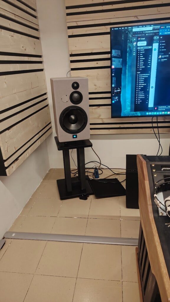 Mum 10 A speaker in the corner of a room on top of a stand with the left corner of a large video monitor showing and 2 pine wood panel sound treatment panels